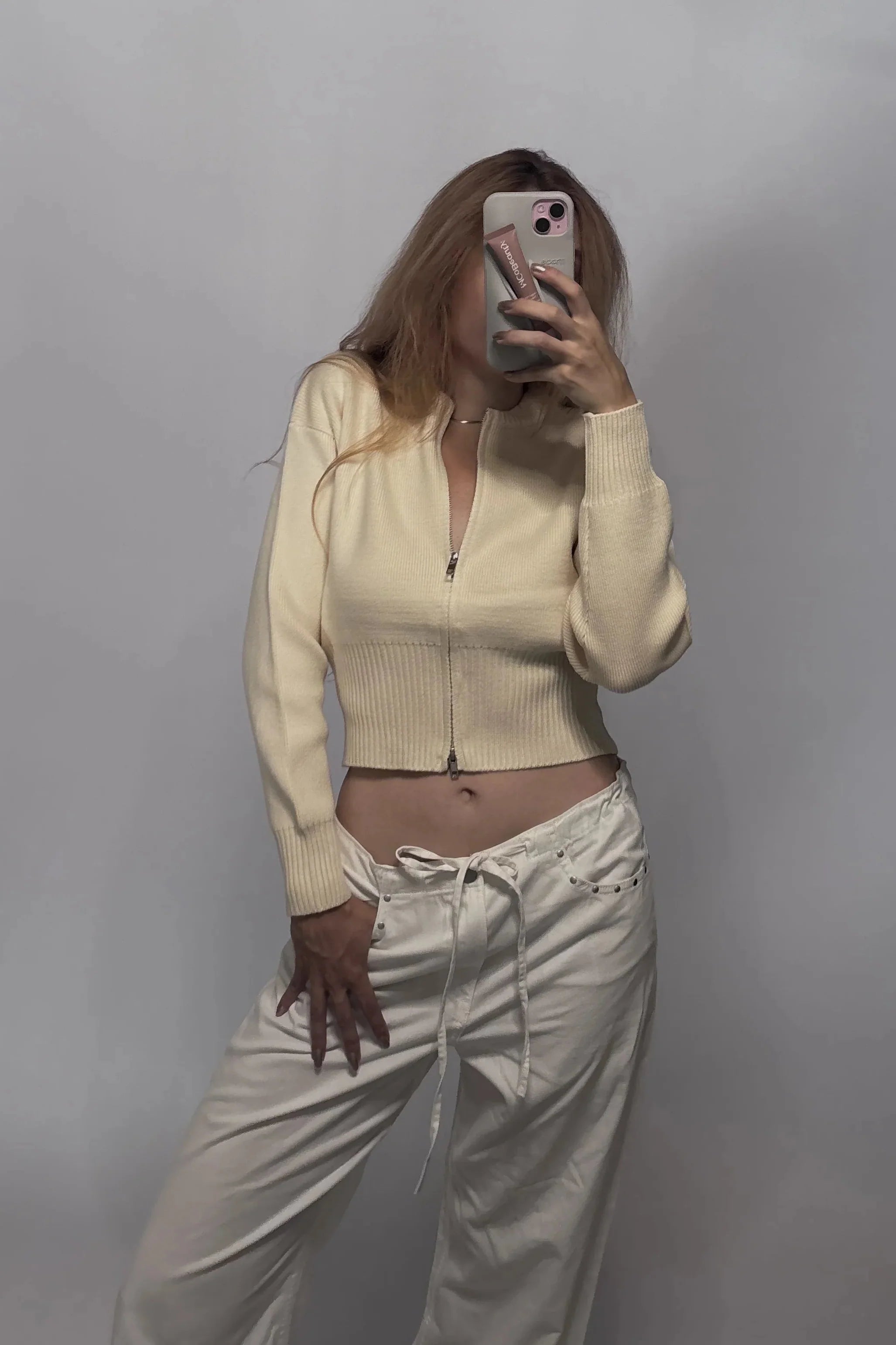A woman takes a mirror selfie wearing a cream-colored ribbed zip-up crop top and matching cream drawstring pants against a plain gray wall.