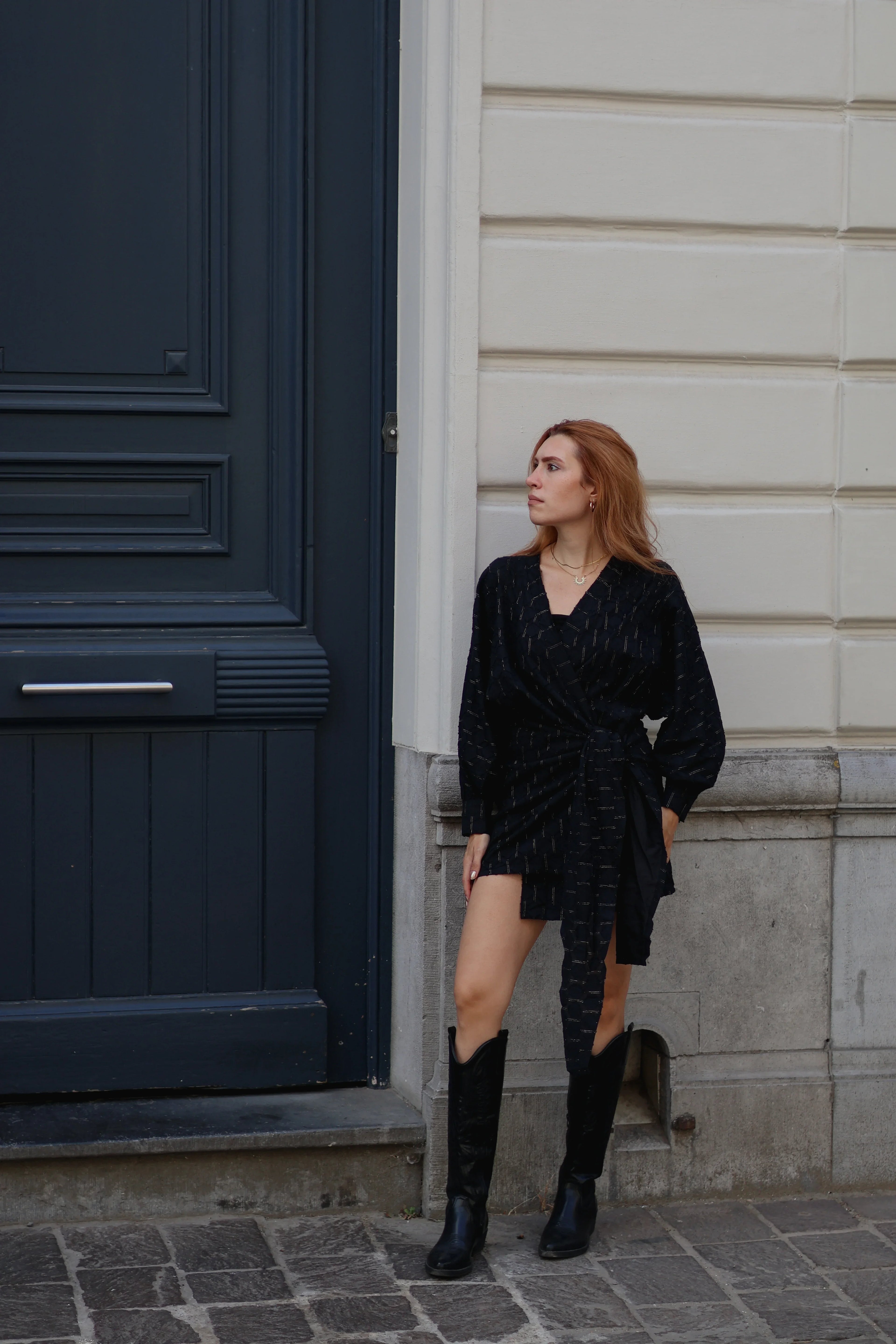 A woman with red hair stands outdoors against a cream-colored wall and a dark blue door, wearing a black textured wrap-style mini dress with long sleeves and a tie waist, paired with black knee-high boots.