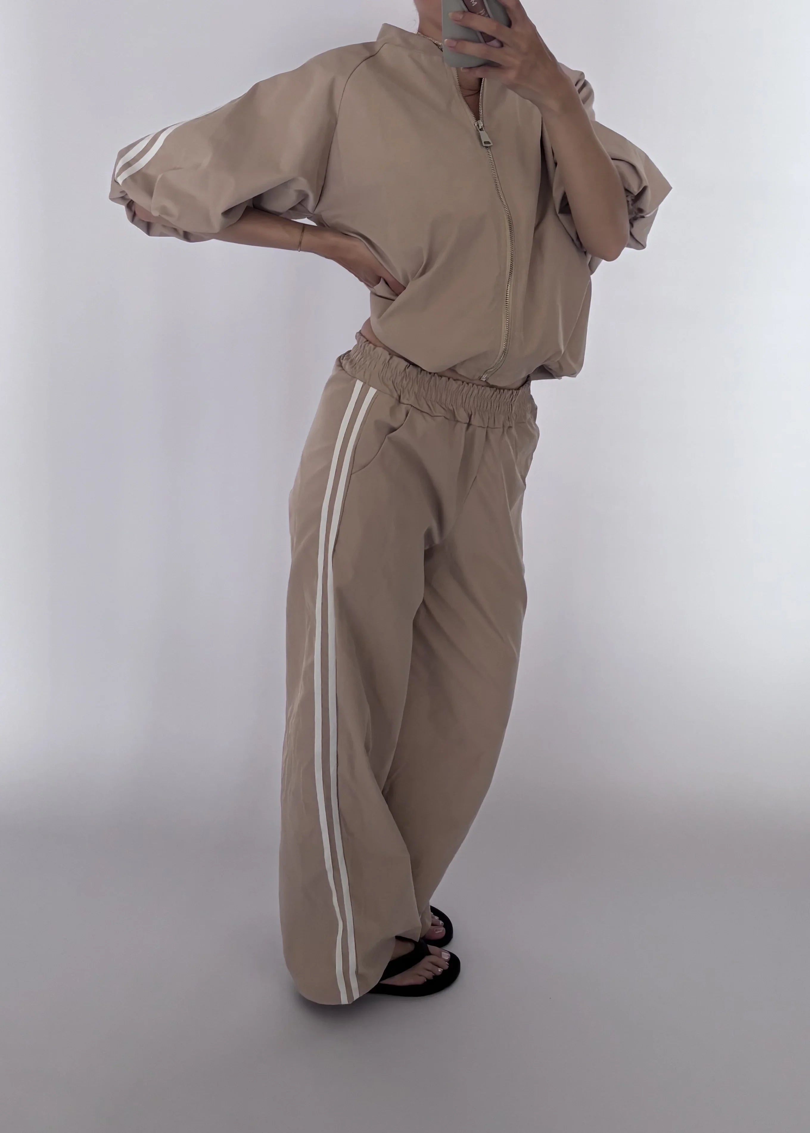 A person modeling a beige zip-up jacket and matching wide-leg sweatpants with white side stripes, photographed against a plain light gray studio background.