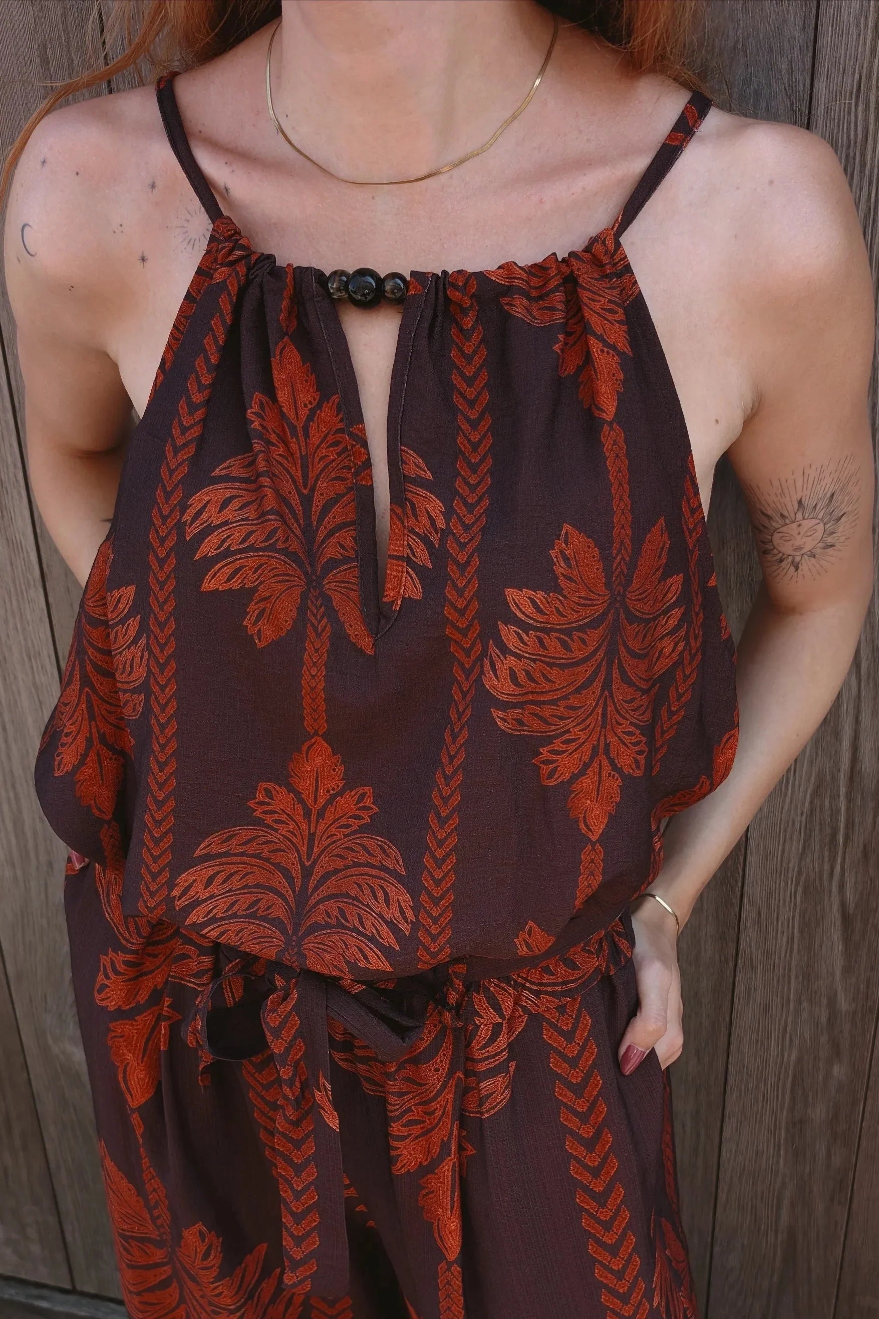A woman wearing a brown and rust-orange patterned halter jumpsuit, standing outdoors against a wooden fence and brick ground.