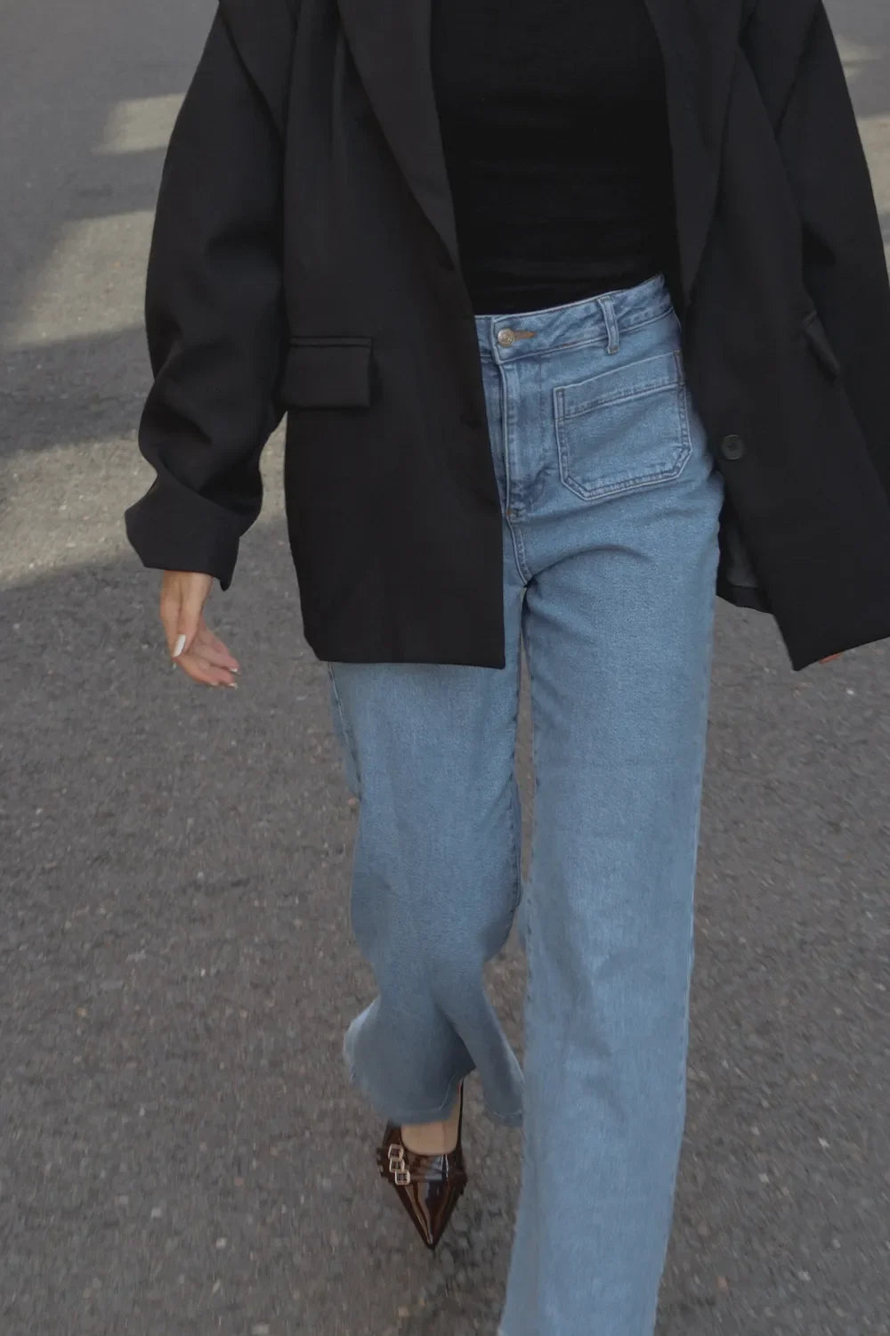 A person walking outdoors wearing a black blazer, black top, light blue wide-leg jeans, and brown pointed shoes on a gray asphalt surface.