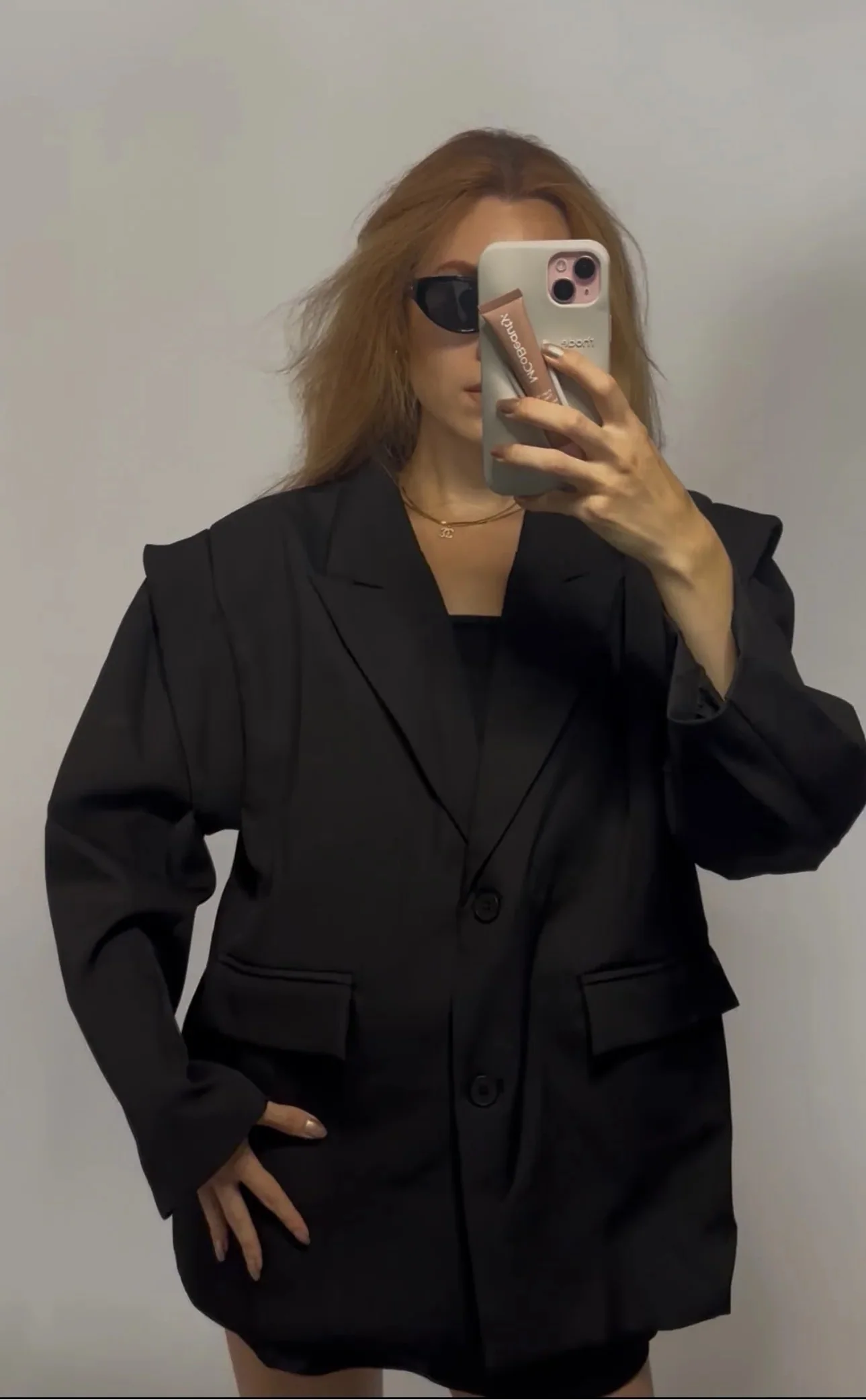 A woman takes a mirror selfie wearing an oversized black blazer with structured shoulders and flap pockets, paired with a black top and black shorts, against a plain light gray background.