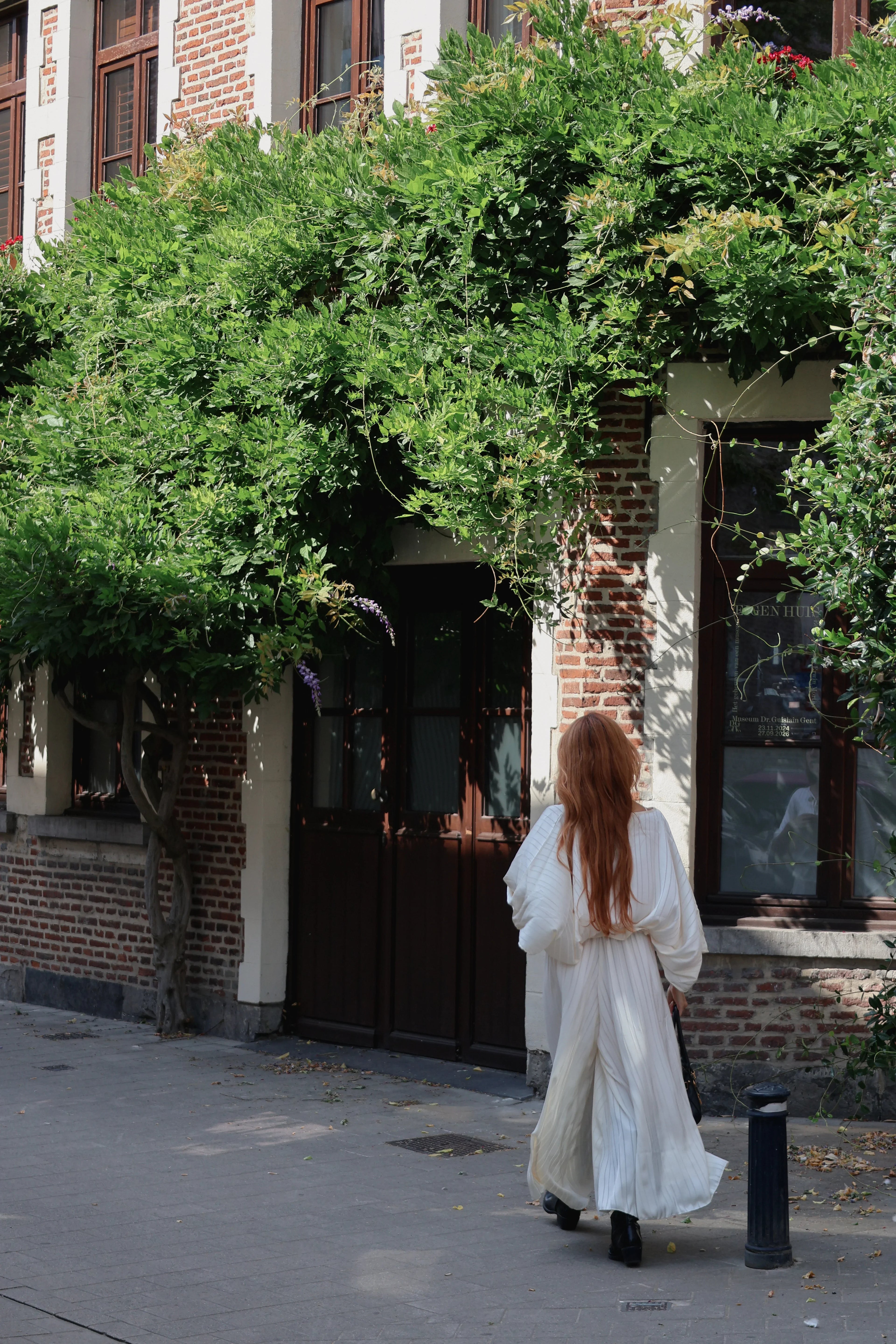 A woman with long red hair wearing a flowing white dress walks away from the camera on a sidewalk in front of a brick building with green vines covering the facade.