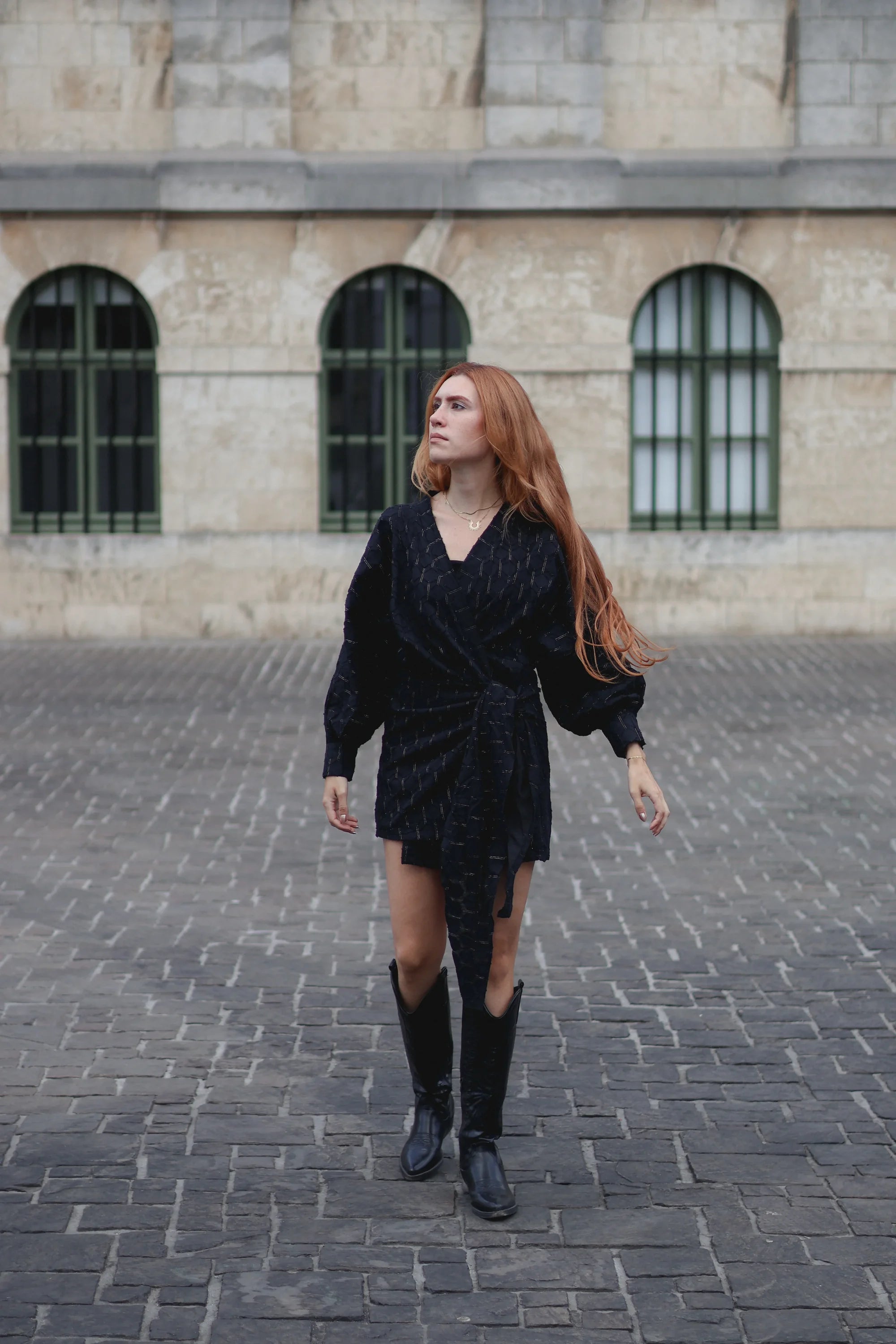 A woman with long red hair wearing a black textured wrap-style mini dress, black knee-high boots, and a gold necklace, walking on a cobblestone street in front of a stone building with arched windows.