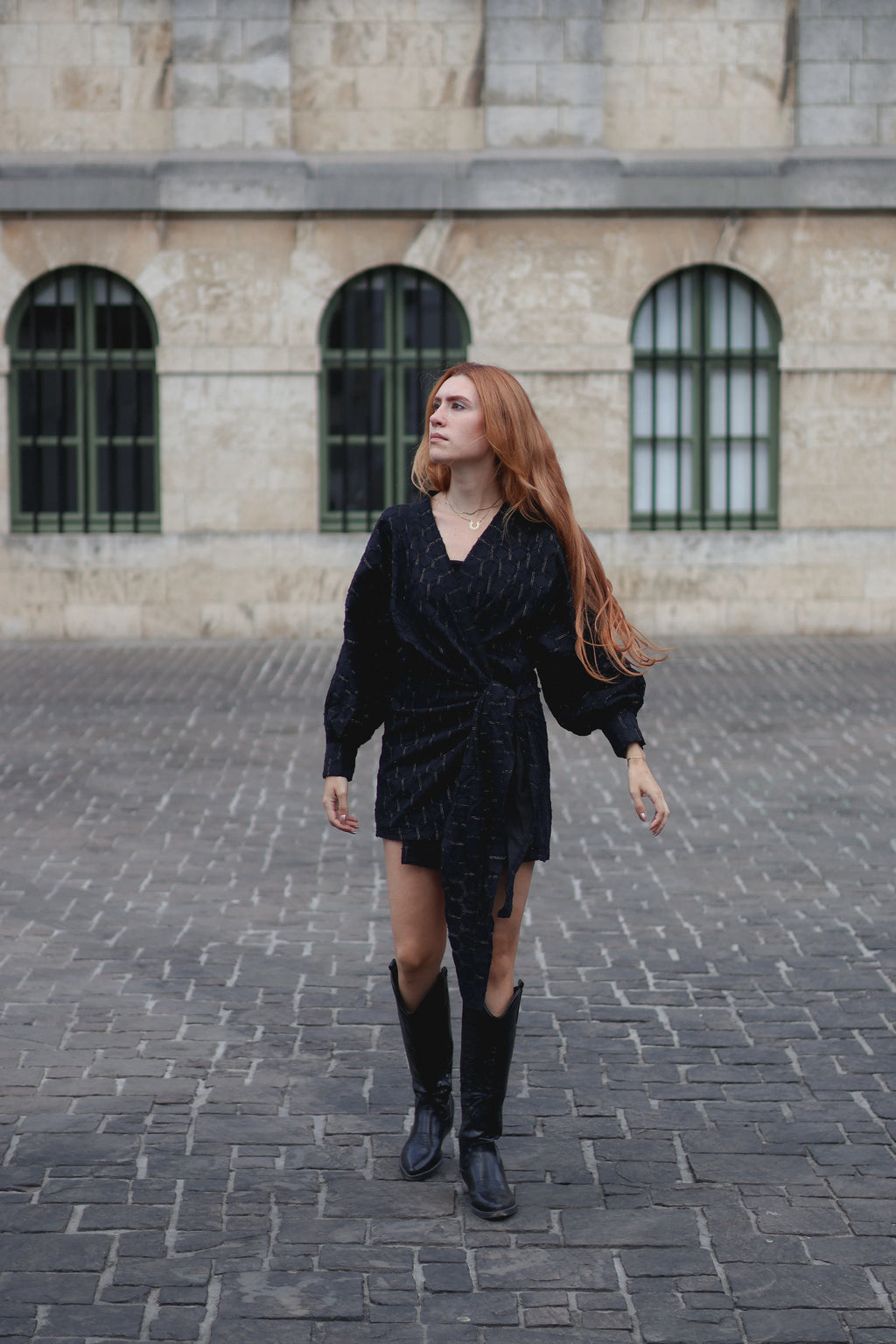 A woman with long red hair wearing a black textured wrap-style mini dress, black knee-high boots, and a gold necklace, walking on a cobblestone street in front of a stone building with arched windows.