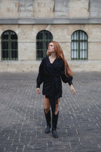 A woman with long red hair wearing a black textured wrap-style mini dress, black knee-high boots, and a gold necklace, walking on a cobblestone street in front of a stone building with arched windows.