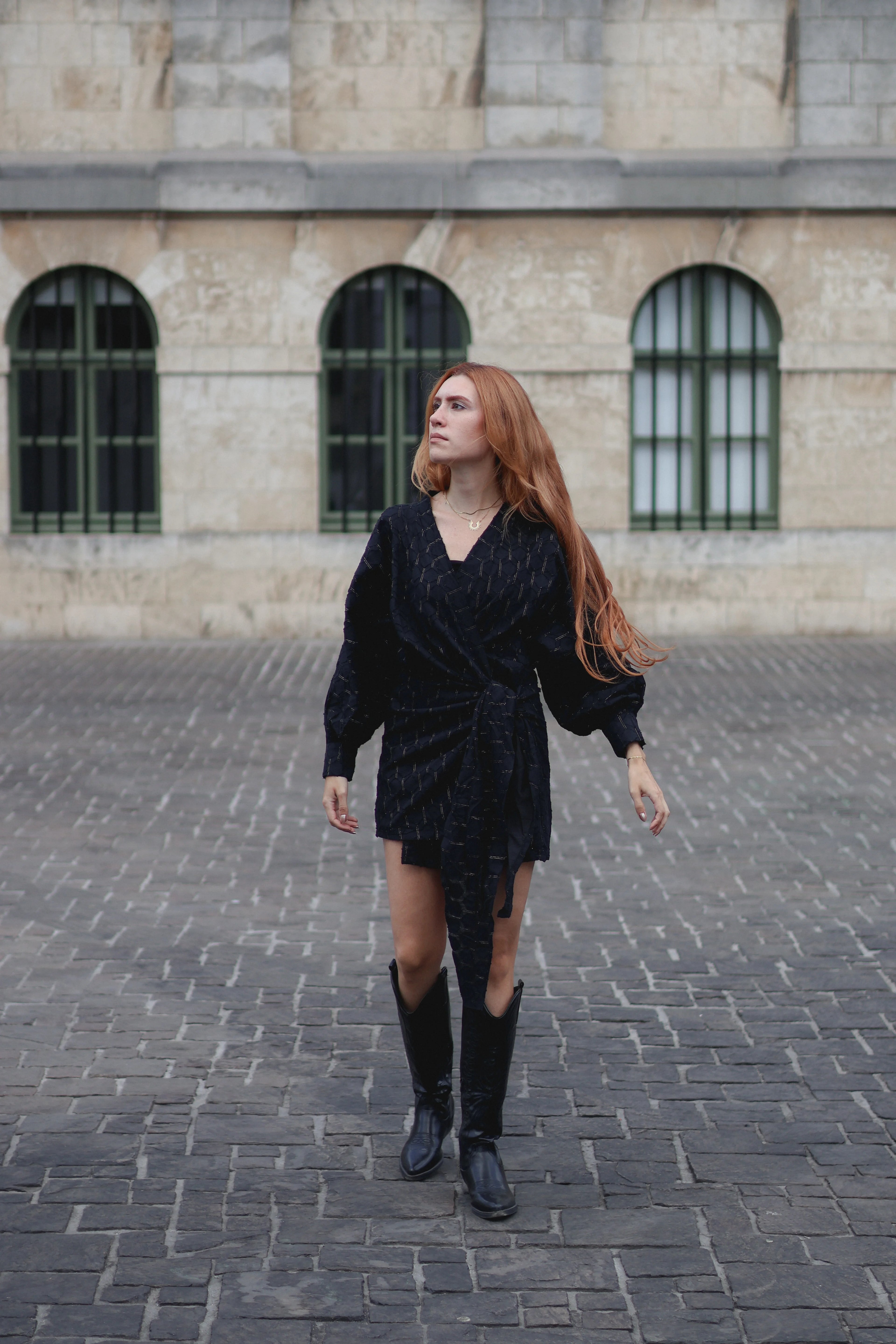 A woman with long red hair wearing a black textured wrap-style mini dress, black knee-high boots, and a gold necklace, walking on a cobblestone street in front of a stone building with arched windows.