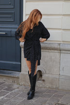 A woman with long strawberry-blonde hair stands outdoors in front of a cream-colored building wall and a dark blue door, wearing a black patterned wrap-style mini dress with long sleeves and black knee-high boots.