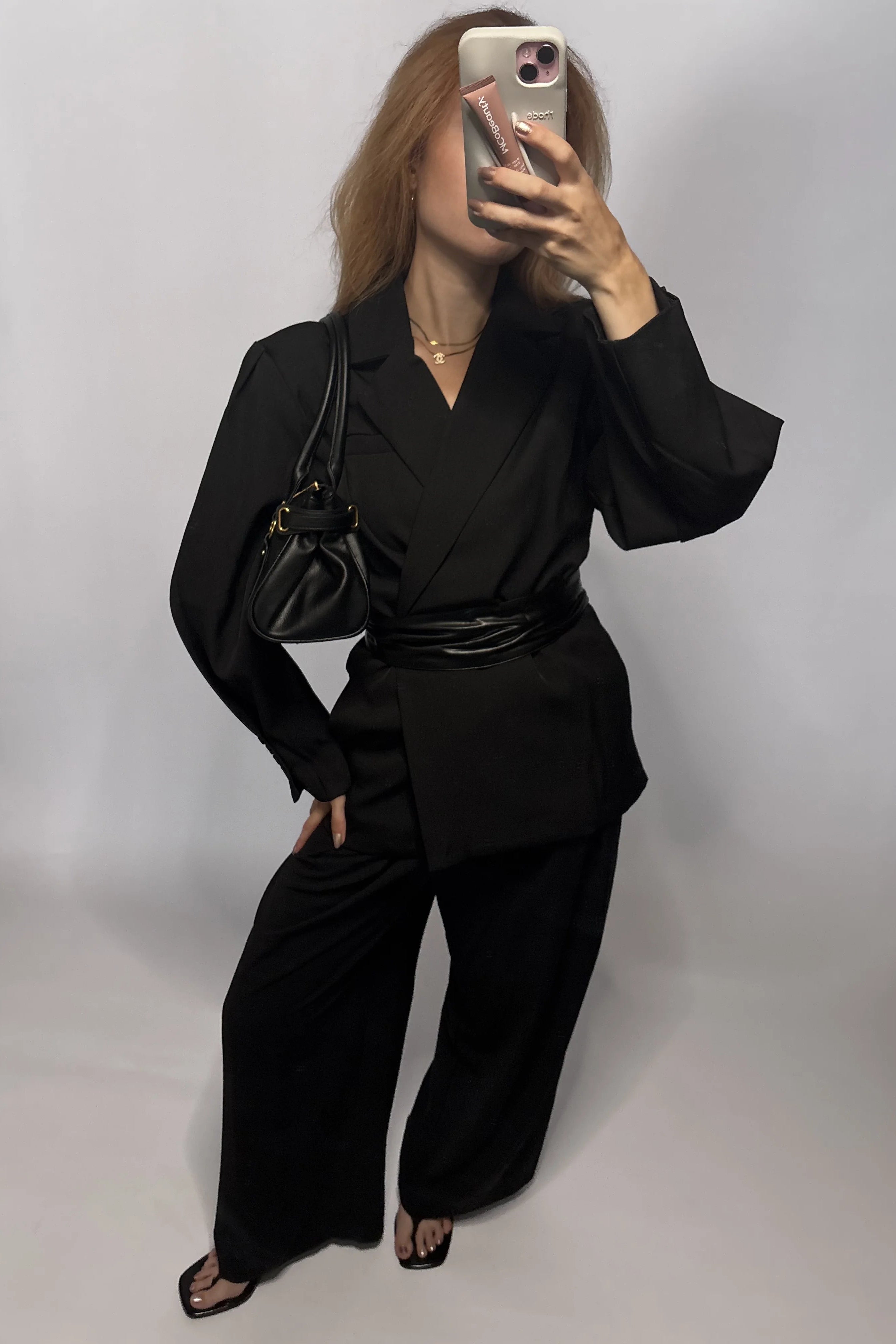 A woman takes a mirror selfie wearing a black tailored blazer with wide-leg matching pants, a black belt, black strappy heels, and carrying a black shoulder bag against a plain light gray studio background.