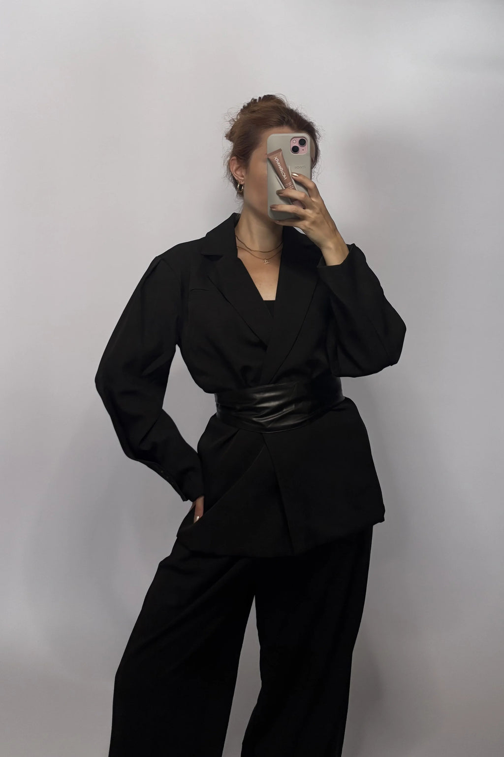 A woman takes a mirror selfie wearing a black long-sleeve blazer-style top with a wide black belt and black wide-leg pants against a plain light gray studio background.
