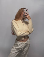 A woman takes a mirror selfie wearing a cream-colored ribbed knit zip-up crop top and matching cream drawstring pants against a plain gray background.