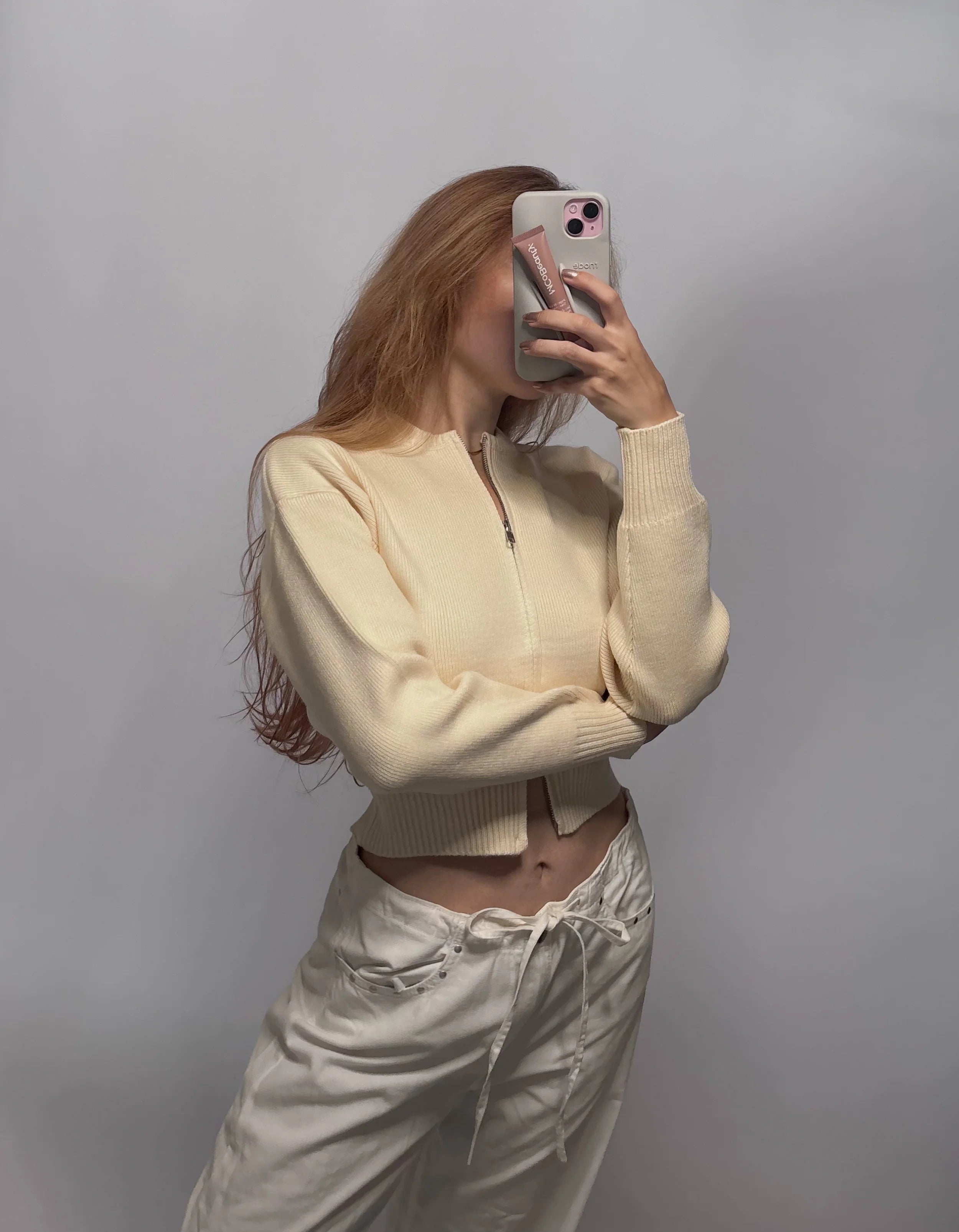 A woman takes a mirror selfie wearing a cream-colored ribbed knit zip-up crop top and matching cream drawstring pants against a plain gray background.