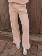 A person wearing light beige wide-leg track pants with white side stripes, paired with sparkly silver sneakers, standing on a brick patio in front of a wooden fence.