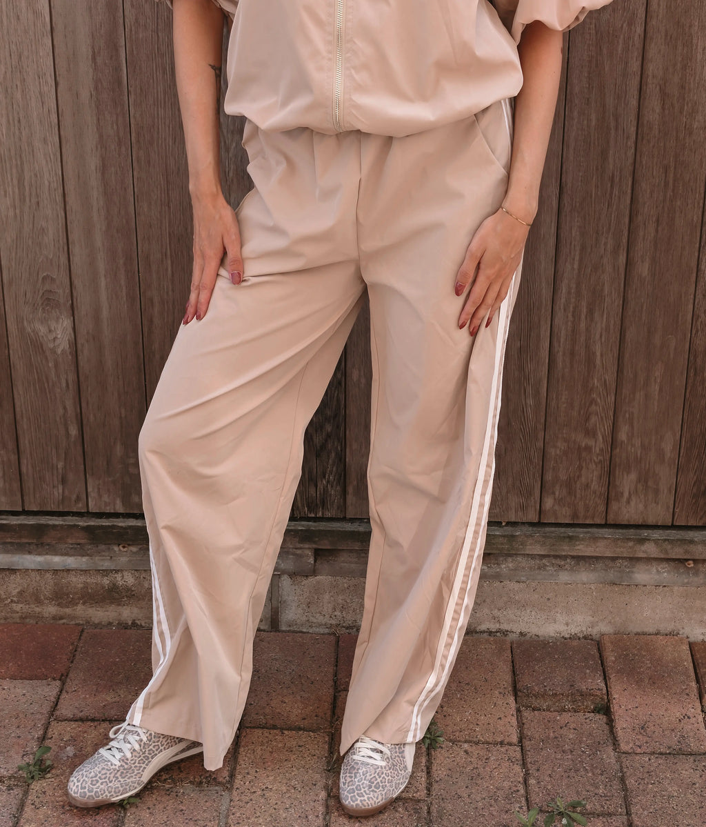 A person wearing loose beige wide-leg pants with white side stripes, paired with a matching beige top and silver glitter sneakers, standing in front of a wooden fence.