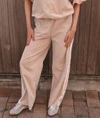A person wearing loose beige wide-leg pants with white side stripes, paired with a matching beige top and silver glitter sneakers, standing in front of a wooden fence.