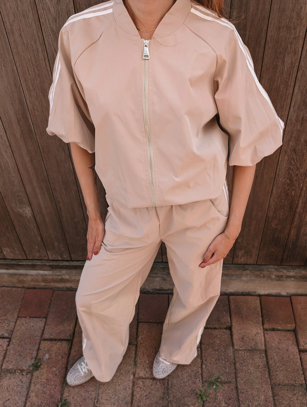 A person wearing a light beige tracksuit with white shoulder stripes, featuring a short-sleeve zip-up jacket and matching wide-leg pants, standing outdoors against a wooden fence.