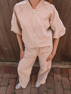 A person wearing a light beige tracksuit with white shoulder stripes, featuring a short-sleeve zip-up jacket and matching wide-leg pants, standing outdoors against a wooden fence.