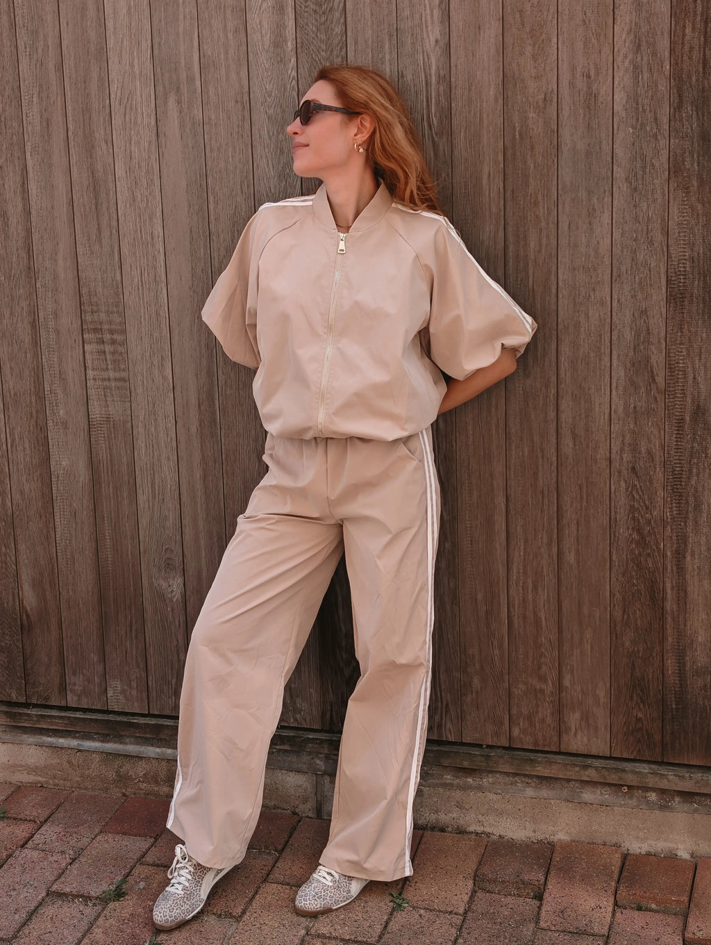 A woman wearing a beige two-piece tracksuit with white side stripes, consisting of a short-sleeve zip-up jacket and matching wide-leg pants, posing against a wooden fence.