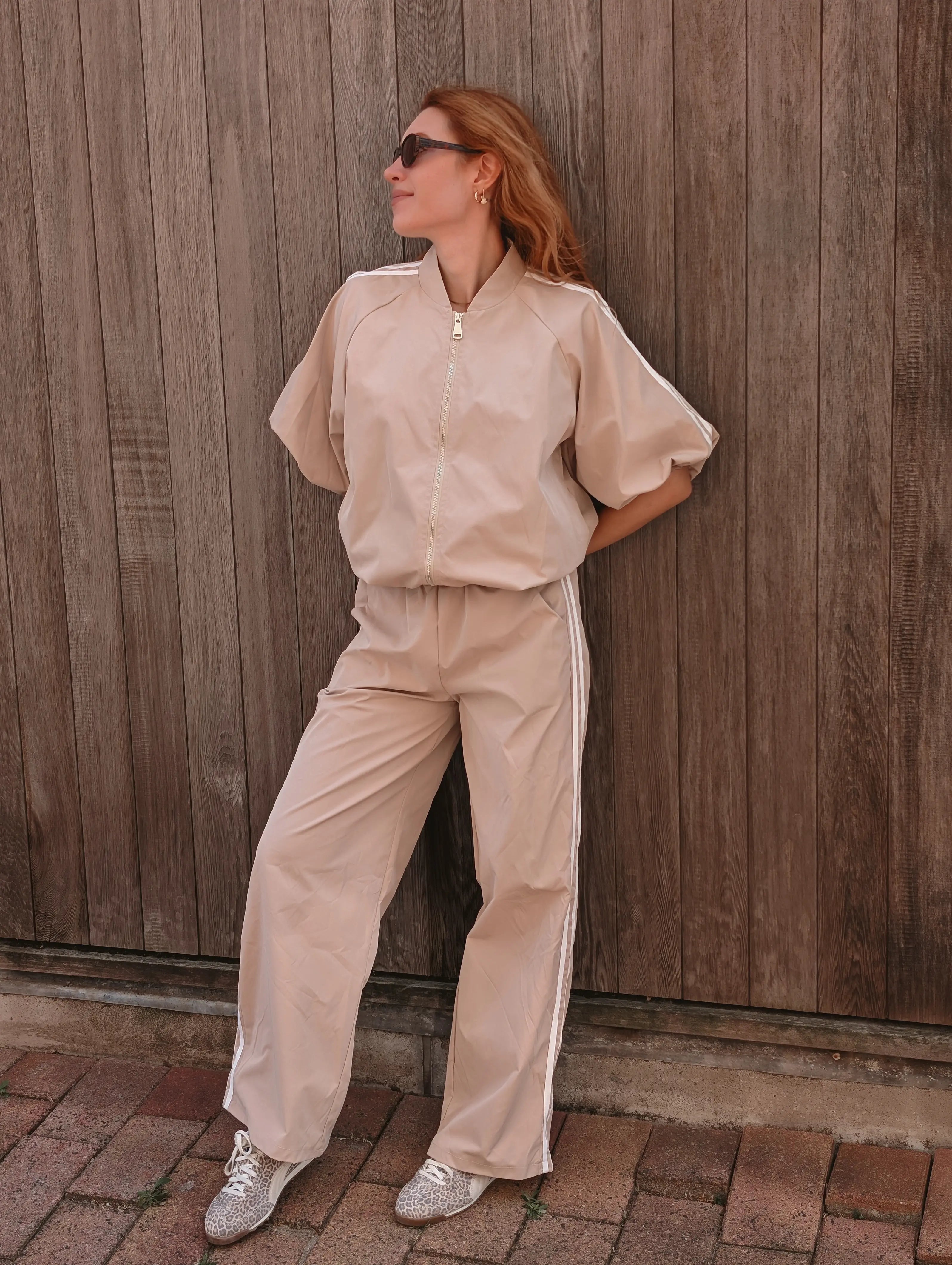 A woman wearing a beige two-piece tracksuit with white side stripes, consisting of a short-sleeve zip-up jacket and matching wide-leg pants, posing against a wooden fence.