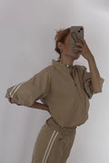 A person taking a mirror selfie wearing a beige matching two-piece tracksuit with white side stripes, consisting of a loose long-sleeve top and wide-leg pants, paired with black sandals.