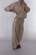 A person modeling a beige zip-up jacket and matching wide-leg sweatpants with white side stripes, photographed against a plain light gray studio background.