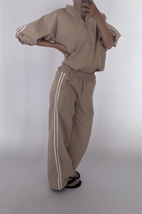 A person modeling a beige zip-up jacket and matching wide-leg sweatpants with white side stripes, photographed against a plain light gray studio background.