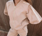 A person wearing a light beige tracksuit with white double stripes down the sleeves and legs, featuring a front zipper, photographed against a dark wooden plank background.