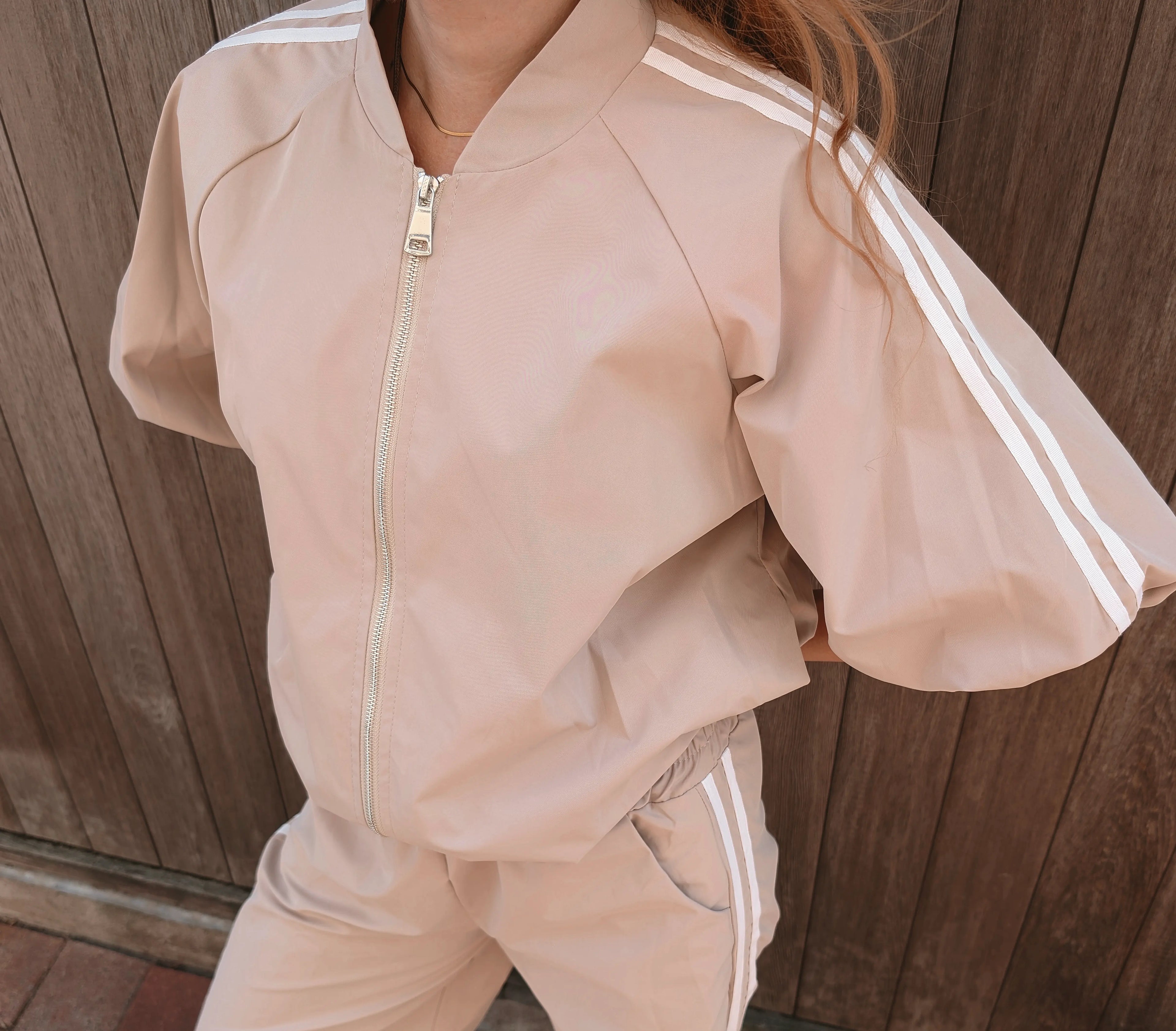 A person wearing a light beige tracksuit with white double stripes down the sleeves and legs, featuring a front zipper, photographed against a dark wooden plank background.