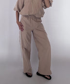 A model wearing beige wide-leg pants with white side stripes, paired with a matching beige top and black sandals, photographed against a light gray studio background.