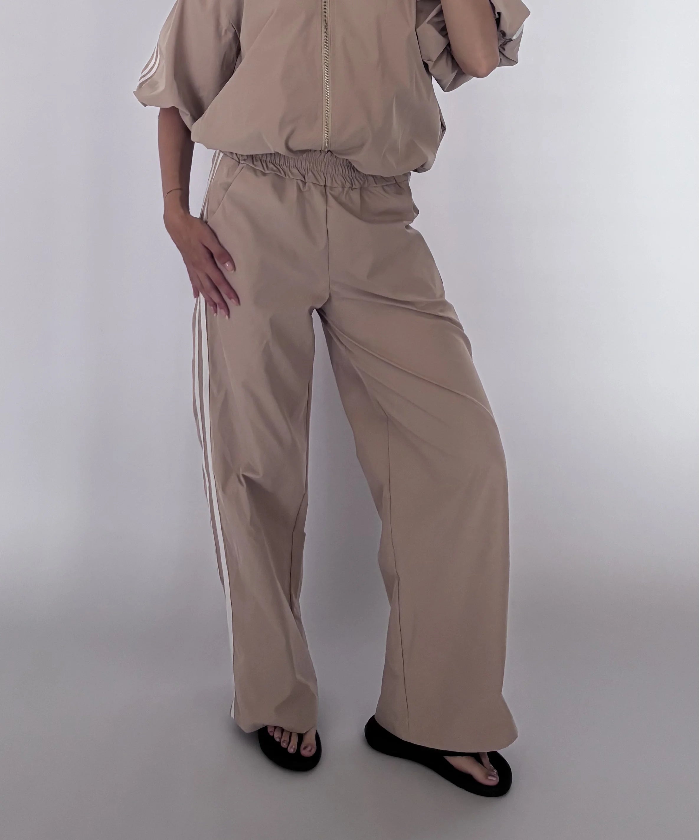 A model wearing beige wide-leg pants with white side stripes, paired with a matching beige top and black sandals, photographed against a light gray studio background.