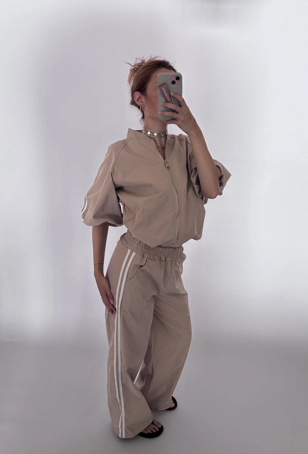 A woman takes a mirror selfie wearing a matching beige two-piece outfit consisting of a short-sleeve zip-up jacket and wide-leg pants with white side stripes, set against a plain light gray background.