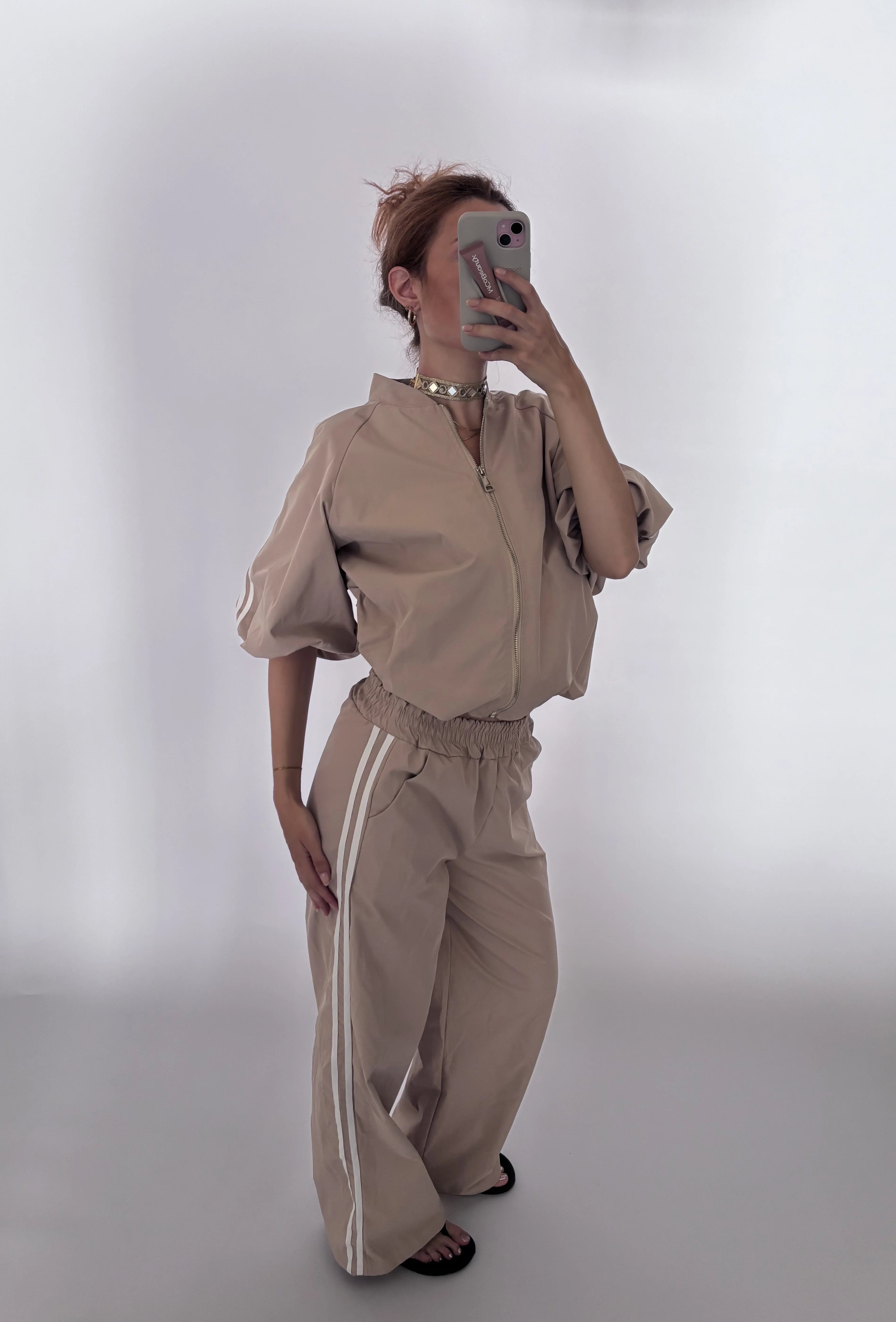 A woman takes a mirror selfie wearing a matching beige two-piece outfit consisting of a short-sleeve zip-up jacket and wide-leg pants with white side stripes, set against a plain light gray background.