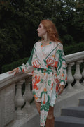 A woman with red hair stands on outdoor stone stairs wearing a long-sleeve wrap-style dress with a cream base and a bold botanical pattern in green, orange, and white. Trees are visible in the background.