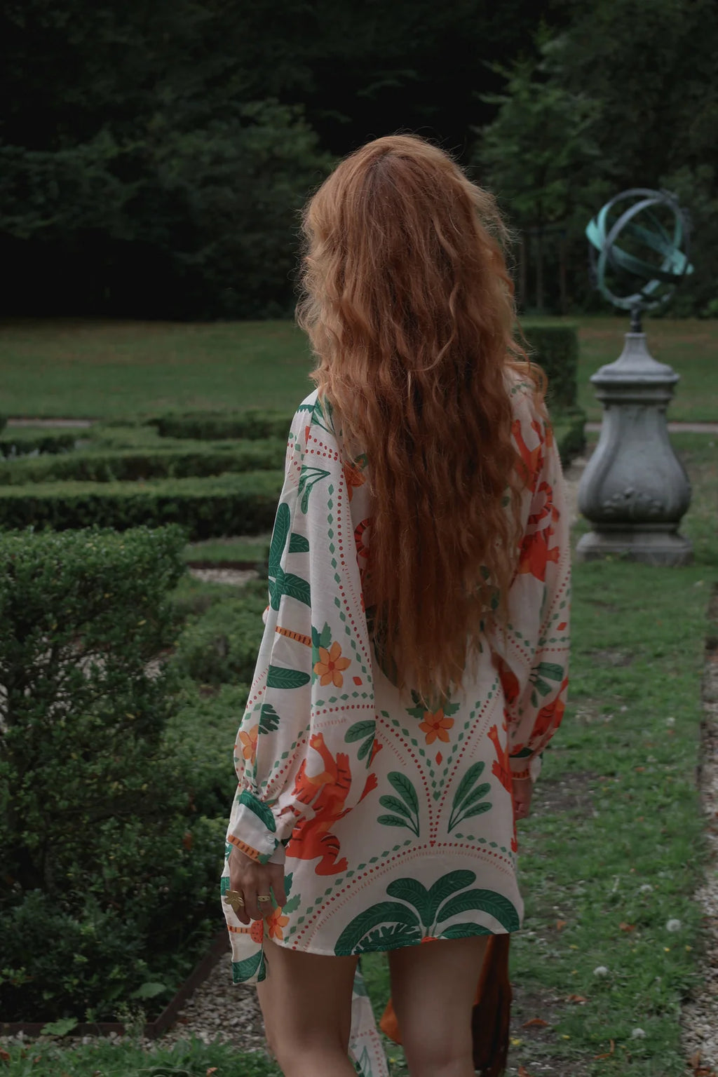 A woman with long, wavy strawberry-blonde hair is seen from behind, wearing a bohemian-style printed kimono cover-up. She is standing in a formal garden with trimmed hedges, a stone armillary sphere, and dark green trees in the background.