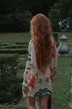 A woman with long, wavy strawberry-blonde hair is seen from behind, wearing a bohemian-style printed kimono cover-up. She is standing in a formal garden with trimmed hedges, a stone armillary sphere, and dark green trees in the background.