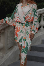 A woman wearing a cream wrap-style dress with a green and orange botanical print, tied at the waist, standing on outdoor stone steps.