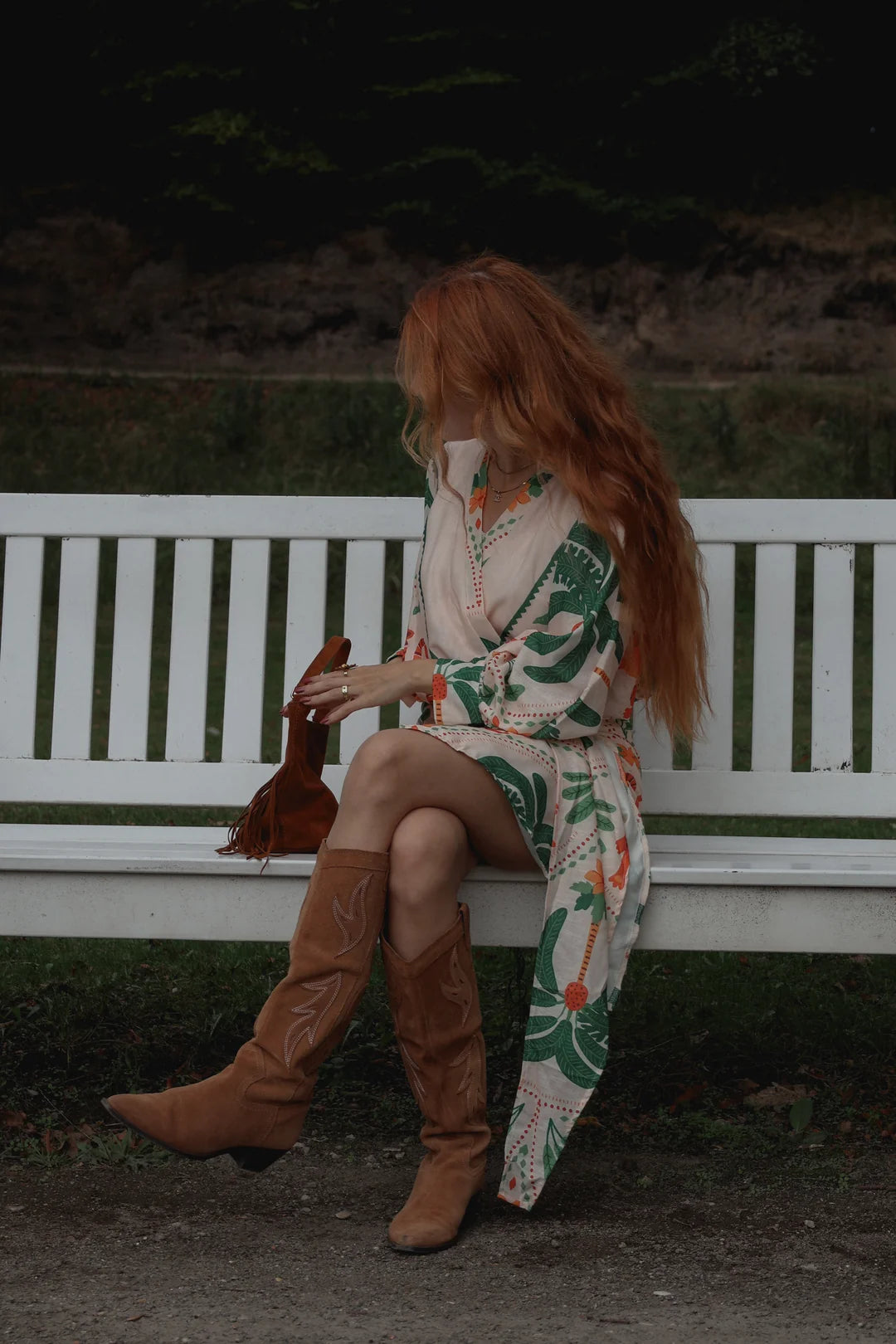 A woman with long red hair sits on a white wooden bench outdoors, wearing a cream boho-style dress with green and orange floral patterns, brown cowboy boots, and holding a brown leather bag.