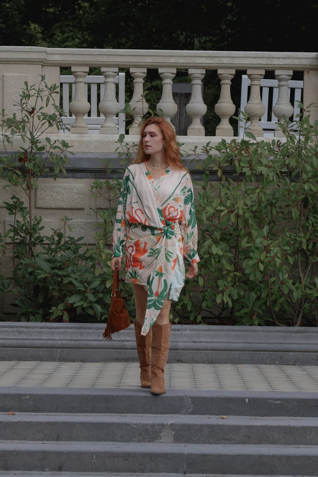 A woman with red hair stands on outdoor stone steps wearing a cream wrap-style dress with a green and orange floral print, paired with brown suede boots and holding a brown handbag.