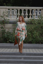 A woman with red hair stands on outdoor stone steps wearing a cream wrap-style dress with a green and orange floral print, paired with brown suede boots and holding a brown handbag.