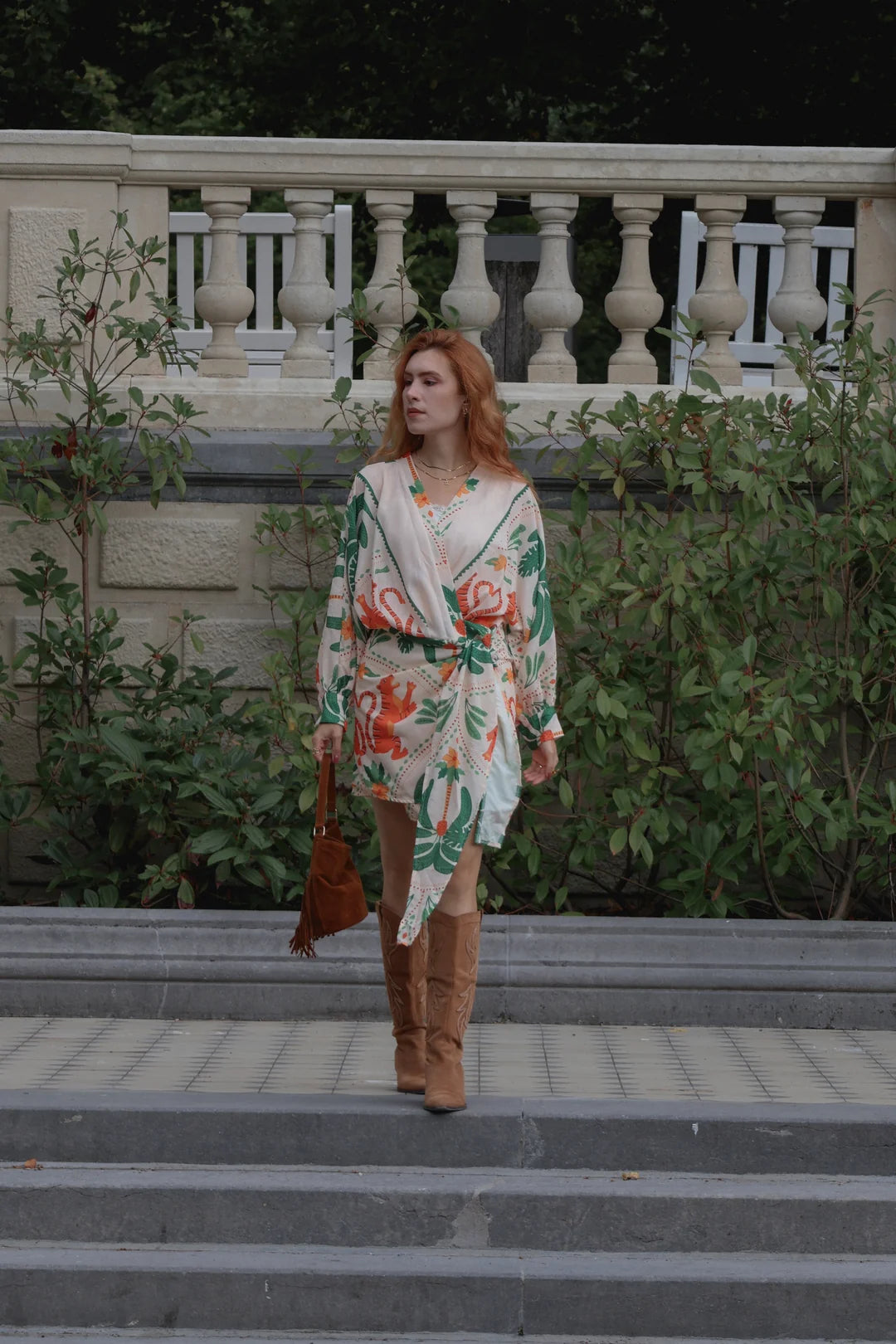 A woman with red hair stands on outdoor stone steps wearing a cream wrap-style dress with a green and orange floral print, paired with brown suede boots and holding a brown handbag.