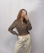 A woman takes a mirror selfie wearing a cropped, long-sleeve zip-up knit top in taupe brown and cream wide-leg pants against a plain light gray background.