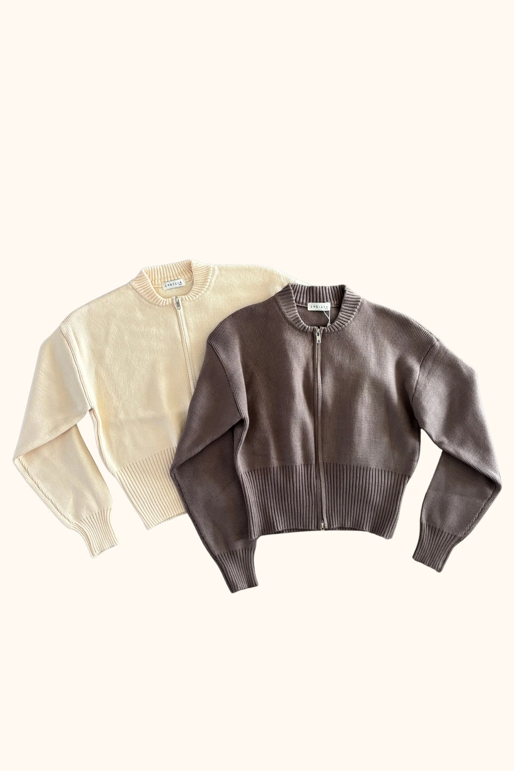 Two cropped zip-up knit cardigans laid flat on a white background, one in cream and one in taupe-brown. The front cardigan is brown and overlaps the cream one behind it.