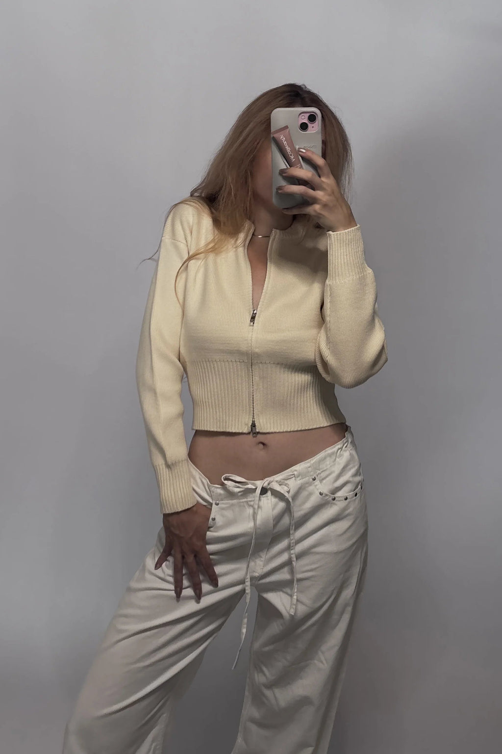 A woman takes a mirror selfie wearing a cream-colored ribbed zip-up crop top and matching cream drawstring pants against a plain gray wall.