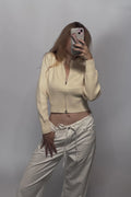 A woman takes a mirror selfie wearing a cream-colored ribbed zip-up crop top and matching cream drawstring pants against a plain gray wall.