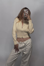 A woman takes a mirror selfie wearing a cream-colored ribbed zip-up crop top and matching cream drawstring pants against a plain gray wall.