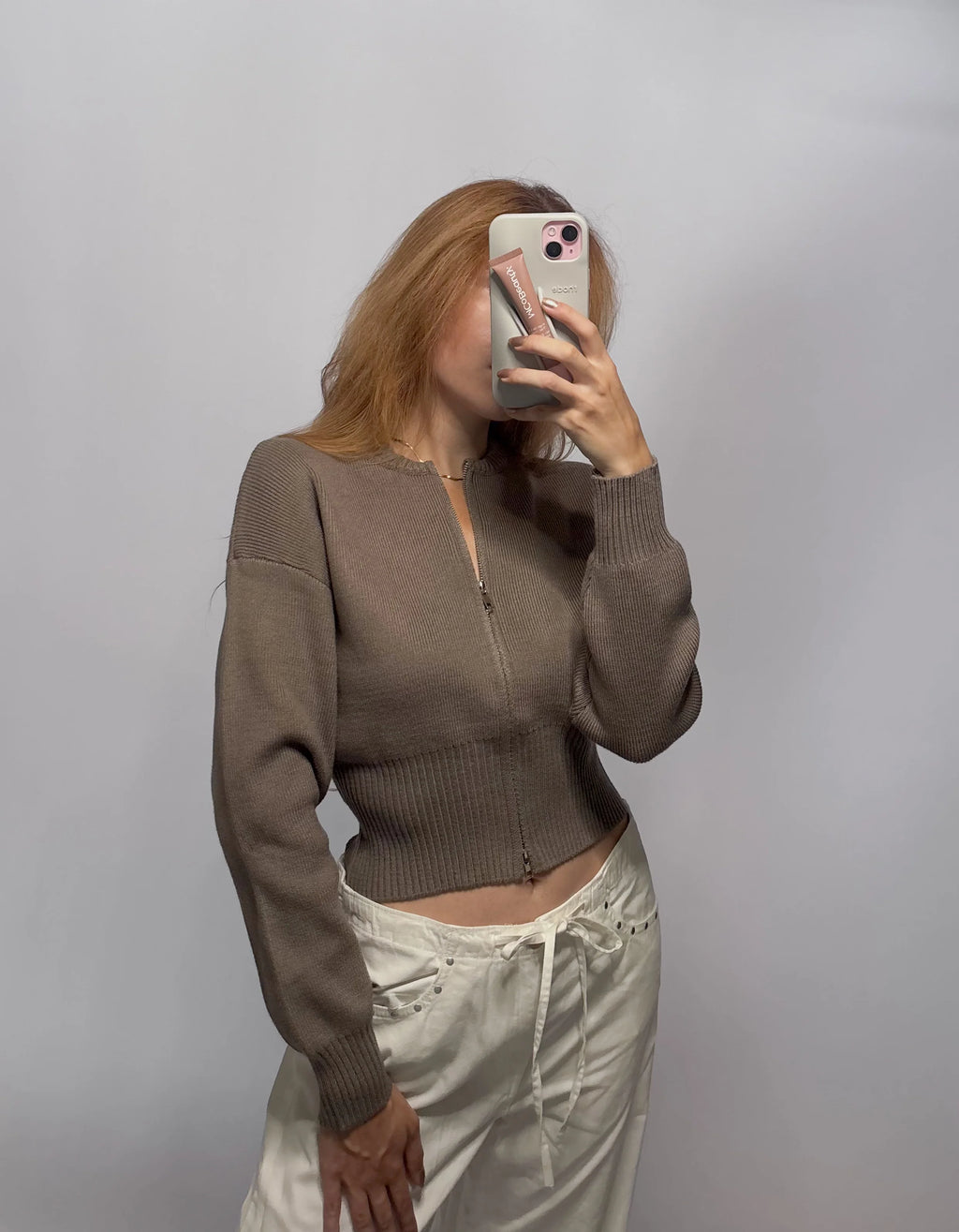 A woman takes a mirror selfie wearing a cropped taupe-brown ribbed knit zip-up cardigan and cream-colored drawstring pants against a plain gray studio background.