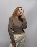 A woman takes a mirror selfie wearing a cropped taupe-brown ribbed knit zip-up cardigan and cream-colored drawstring pants against a plain gray studio background.