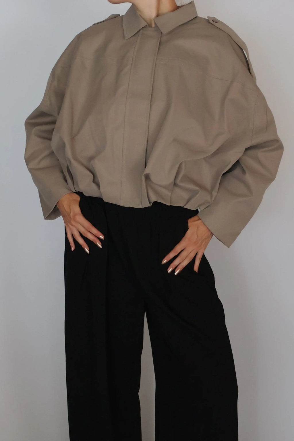 A woman with red hair tied up is wearing a loose taupe-brown long-sleeve button-up shirt and black wide-leg trousers, standing against a plain light gray studio background.