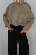A woman with red hair tied up is wearing a loose taupe-brown long-sleeve button-up shirt and black wide-leg trousers, standing against a plain light gray studio background.