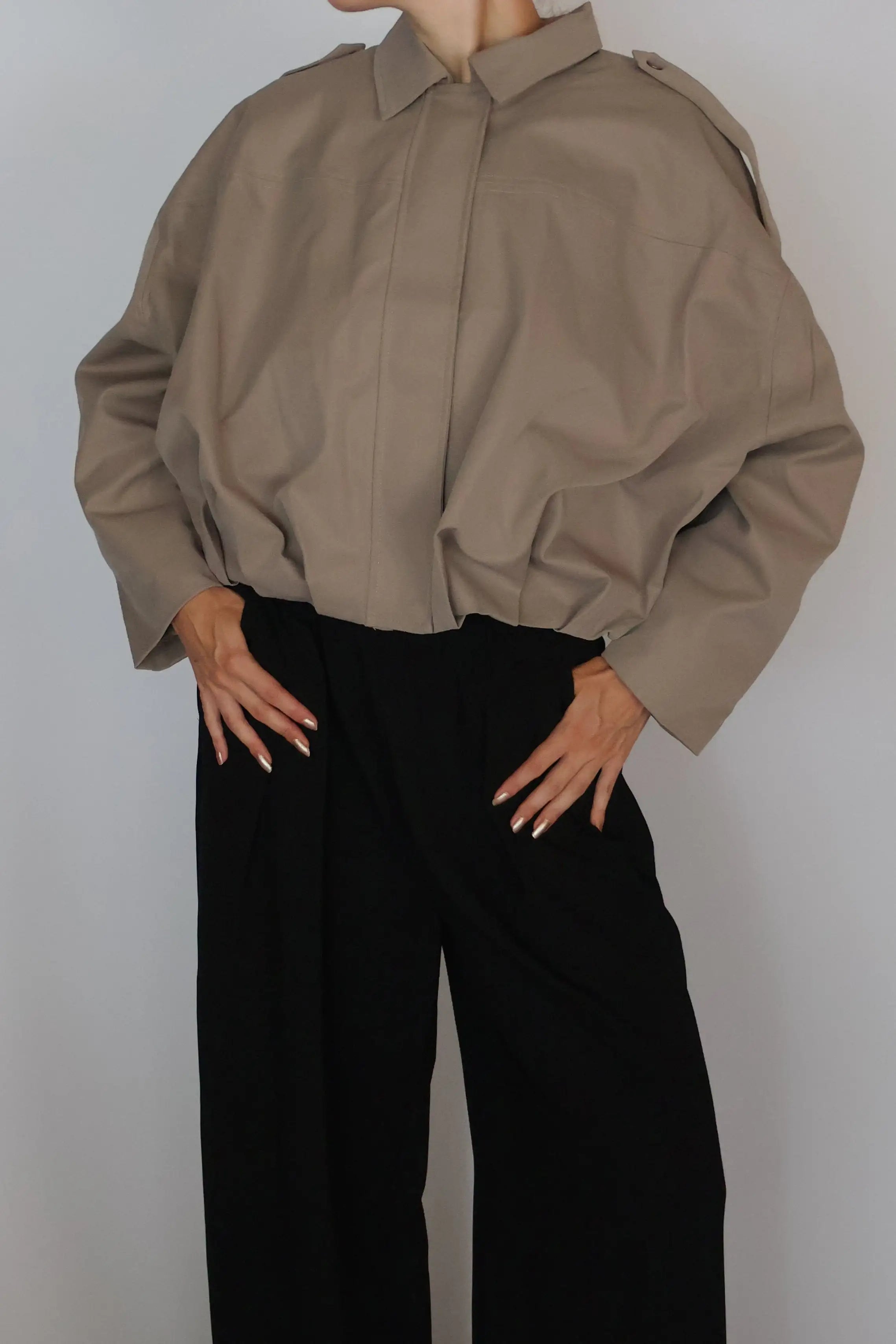 A woman with red hair tied up is wearing a loose taupe-brown long-sleeve button-up shirt and black wide-leg trousers, standing against a plain light gray studio background.